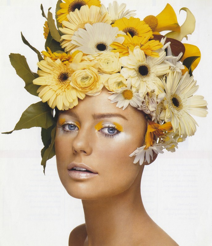 Close-up portrait of a woman with fair skin and blue eyes, wearing elaborate yellow floral headwear and matching yellow eyeshadow in a studio.