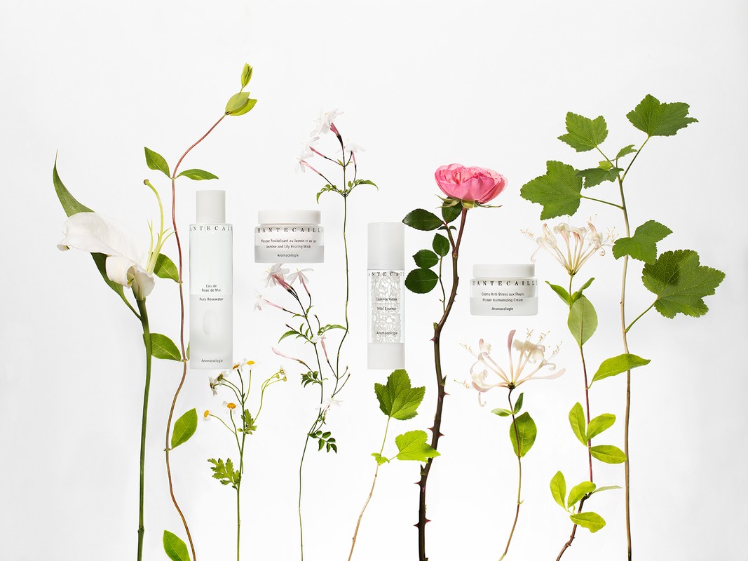 A collection of Chantecaille skincare products is displayed among lush flowers and greenery against a clean, white backdrop in a beauty still life.