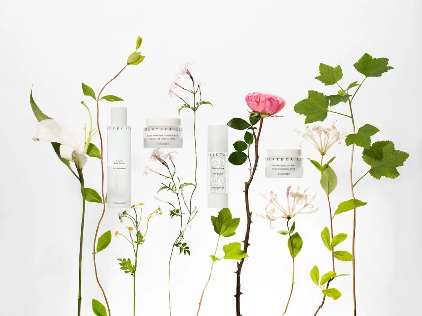 A collection of Chantecaille skincare products is displayed among lush flowers and greenery against a clean, white backdrop in a beauty still life.