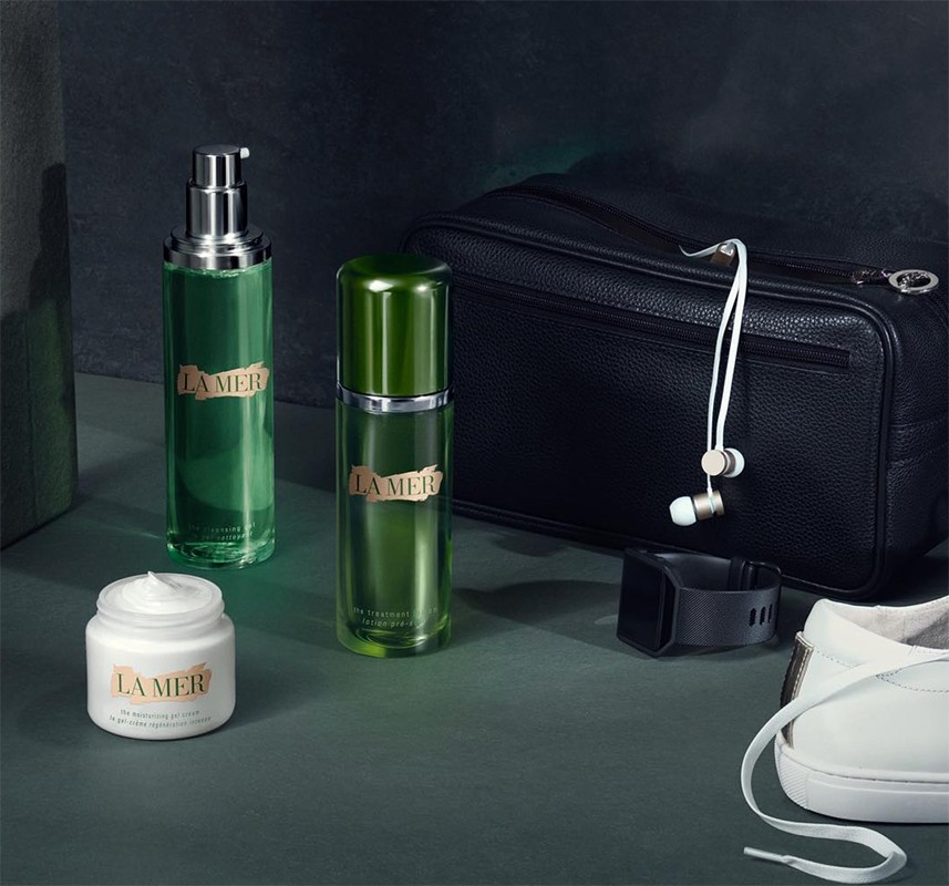 Still life of La Mer skincare products including creams, serum, and lotion, along with a black travel bag, smart watch, and white sneakers.