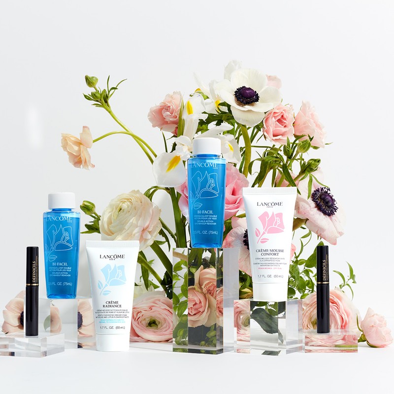 Lancôme skincare and mascara products arranged on clear display blocks against a backdrop of roses and anemones on a white background.