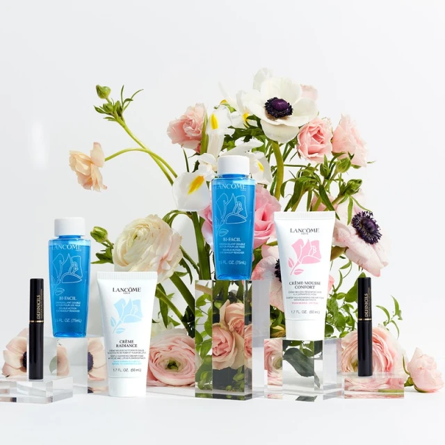 Lancôme skincare and mascara products arranged on clear display blocks against a backdrop of roses and anemones on a white background.