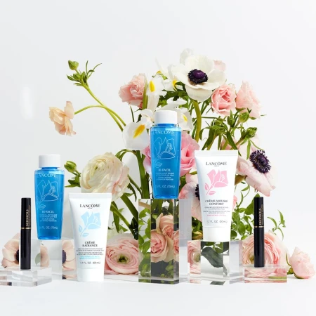 Lancôme skincare and mascara products arranged on clear display blocks against a backdrop of roses and anemones on a white background.