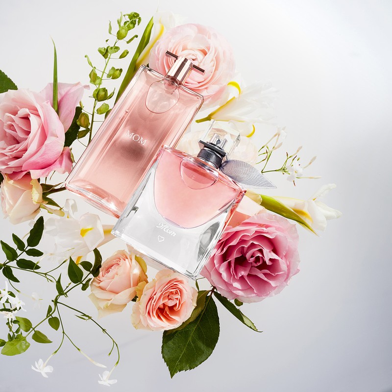 Two perfume bottles, one pink and one clear, surrounded by pink and white roses, lilies, and small white flowers on a white surface.