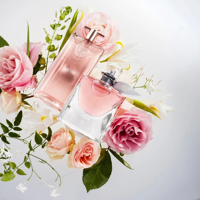 Two perfume bottles, one pink and one clear, surrounded by pink and white roses, lilies, and small white flowers on a white surface.