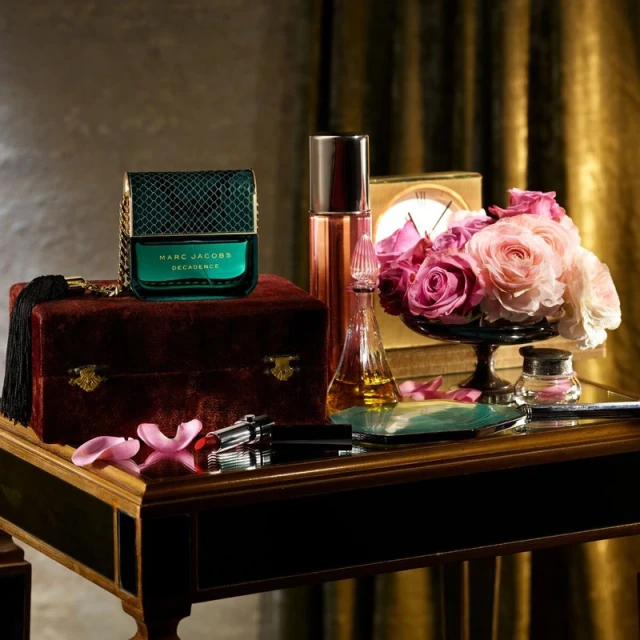 Still life featuring perfume bottles, roses, and vanity items arranged on a polished table against a draped, golden background.