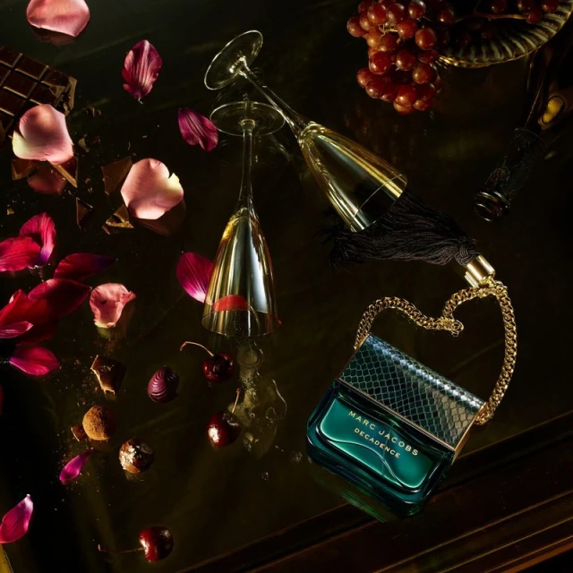 Elevated view of Marc Jacobs Decadence perfume bottle with gold chain, cherries, rose petals, chocolate, grapes, and champagne flutes.