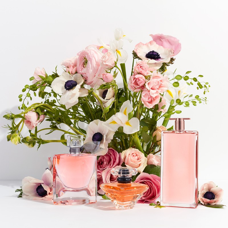 Three perfume bottles are arranged in front of a lush bouquet of pink and white flowers, with a bright white background and studio lighting.