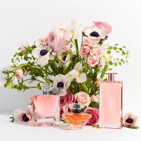 Three perfume bottles are arranged in front of a lush bouquet of pink and white flowers, with a bright white background and studio lighting.