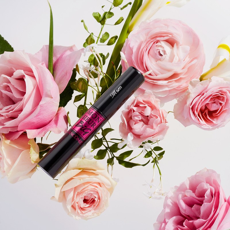 Close-up of a black mascara tube surrounded by pink and white roses and green leaves on a white background, beauty concept.
