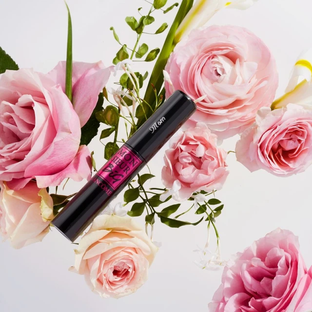 Close-up of a black mascara tube surrounded by pink and white roses and green leaves on a white background, beauty concept.