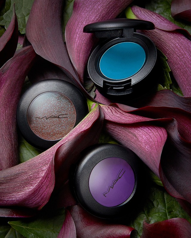 Three MAC eyeshadow compacts in teal, purple, and brown, nestled among vibrant purple calla lilies and green foliage, studio lit.