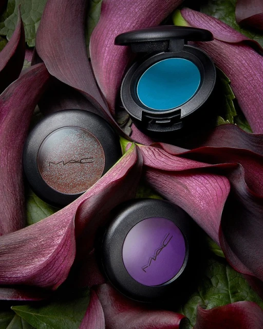 Three MAC eyeshadow compacts in teal, purple, and brown, nestled among vibrant purple calla lilies and green foliage, studio lit.
