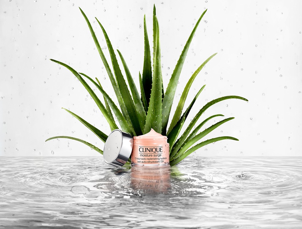 Clinique Moisture Surge skincare product with silver lid surrounded by aloe vera plants, displayed on a reflective water surface.