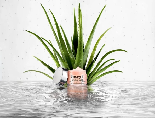 Clinique Moisture Surge skincare product with silver lid surrounded by aloe vera plants, displayed on a reflective water surface.