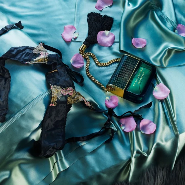A still life composition featuring a designer purse, black lingerie, and scattered pink rose petals on a shiny, silky, turquoise fabric.