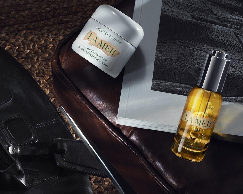 Still life of La Mer skincare products: a jar of cream and a bottle of oil, arranged on a dark leather chair and woven mat.