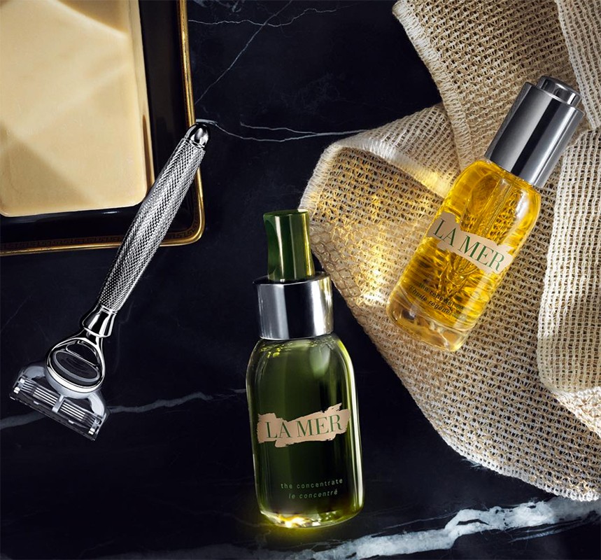 Luxury skincare and grooming product still life featuring La Mer oil and concentrate bottles, a silver razor, and a bar of soap.