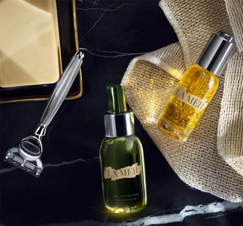 Luxury skincare and grooming product still life featuring La Mer oil and concentrate bottles, a silver razor, and a bar of soap.