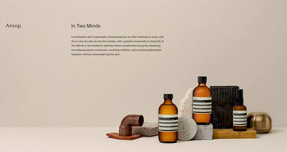 Three Aesop bottles arranged with concrete blocks, terracotta pipe, and gold sphere on a beige surface and background in a studio setting.