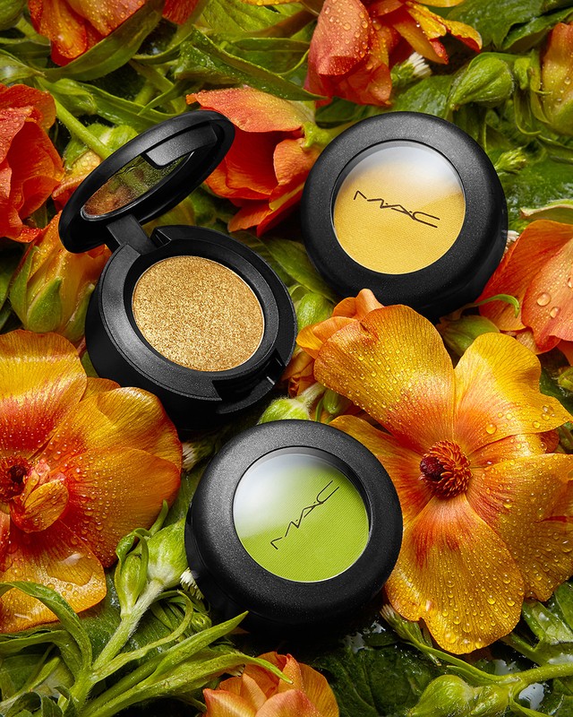 MAC eye shadow pots in yellow, gold, and green hues are displayed among orange flowers and green foliage, set against a dark backdrop.