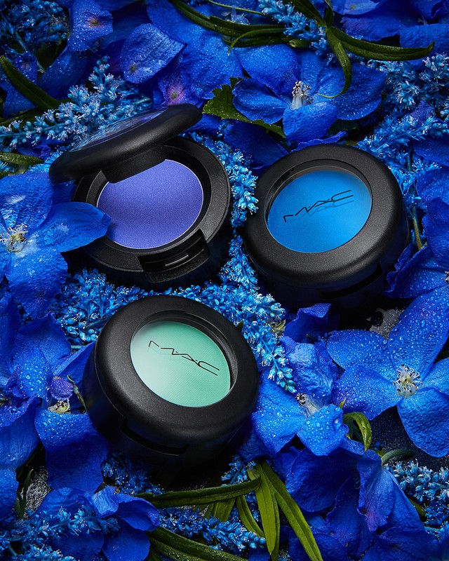 Close-up of three MAC eyeshadows in blue, purple, and green hues nestled among blue flowers with water droplets for a vibrant beauty shot.