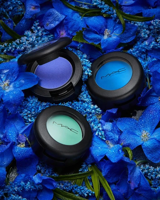 Close-up of three MAC eyeshadows in blue, purple, and green hues nestled among blue flowers with water droplets for a vibrant beauty shot.