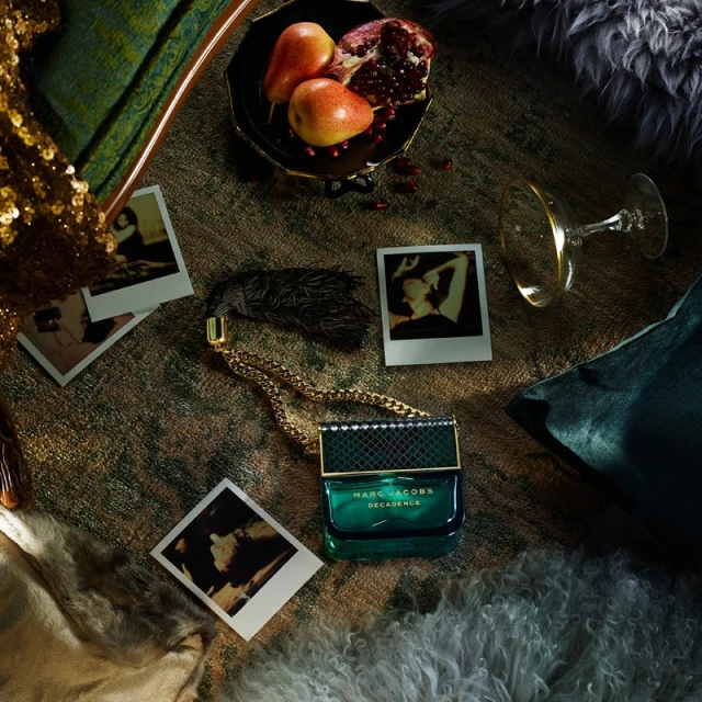 Close-up of Marc Jacobs Decadence perfume with gold chain on patterned surface amid fruit, polaroid photos, and champagne glass.
