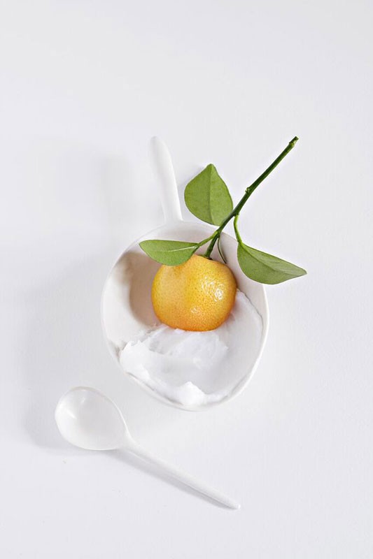 Studio shot of a vibrant yellow-orange tangerine with a leafy green stem on a white spoon, beside a white spoon on a white surface.