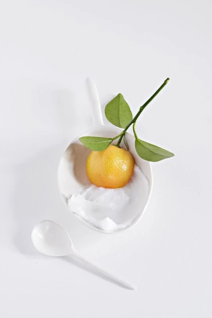 Studio shot of a vibrant yellow-orange tangerine with a leafy green stem on a white spoon, beside a white spoon on a white surface.