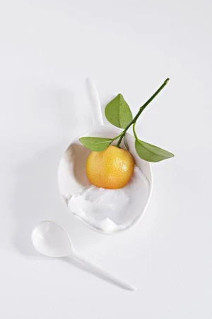 Studio shot of a vibrant yellow-orange tangerine with a leafy green stem on a white spoon, beside a white spoon on a white surface.