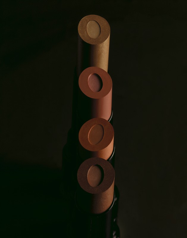 Four lipstick tubes in varying shades of brown are arranged vertically on a dark reflective surface, showcasing makeup and beauty products.