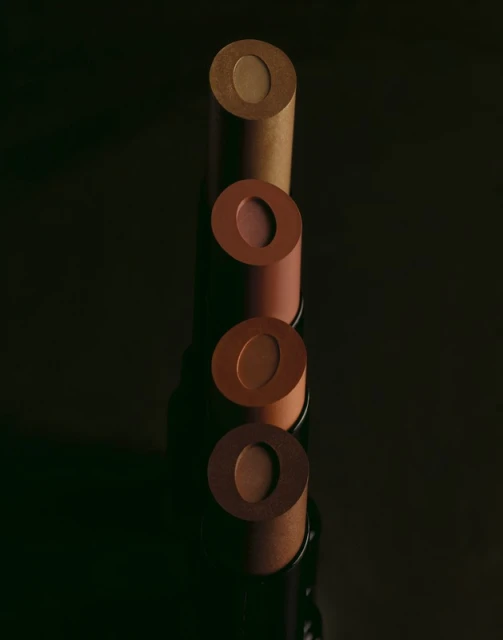 Four lipstick tubes in varying shades of brown are arranged vertically on a dark reflective surface, showcasing makeup and beauty products.
