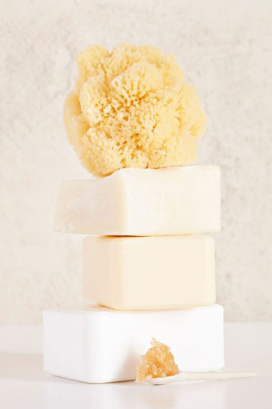 Stacked arrangement of three bars of soap and a natural sponge on a reflective white surface against a textured background.
