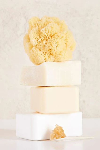 Stacked arrangement of three bars of soap and a natural sponge on a reflective white surface against a textured background.