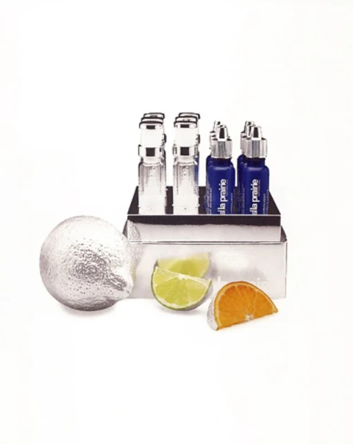 Product still life featuring La Prairie skincare vials in a silver container, alongside silver painted lemon, lime, and orange slices on white.