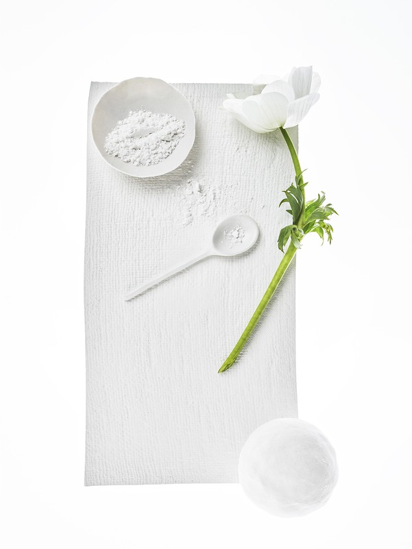 Overhead shot of white anemone flower, powder in bowl, spoon, and sphere on textured white background; monochromatic aesthetic.