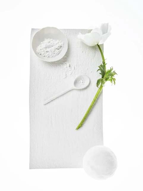 Overhead shot of white anemone flower, powder in bowl, spoon, and sphere on textured white background; monochromatic aesthetic.