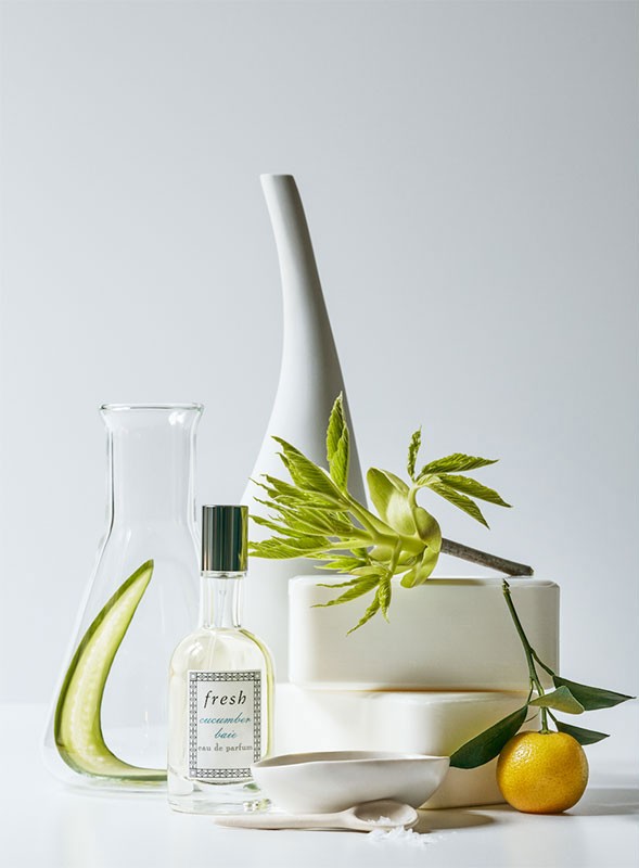 Still life featuring a perfume bottle, laboratory flask, citrus fruit, ceramic bowl, leaves, and bars of soap on a white surface.
