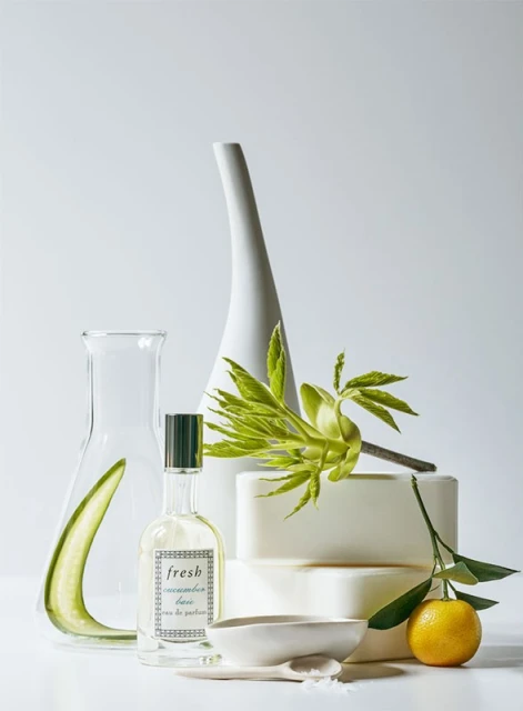 Still life featuring a perfume bottle, laboratory flask, citrus fruit, ceramic bowl, leaves, and bars of soap on a white surface.