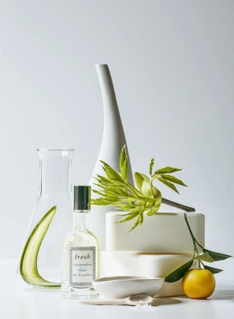 Still life featuring a perfume bottle, laboratory flask, citrus fruit, ceramic bowl, leaves, and bars of soap on a white surface.