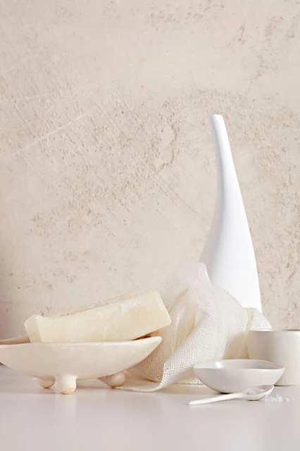 Neutral-toned product shot featuring white soap on a dish next to a white vase and linen, set against a textured off-white backdrop.