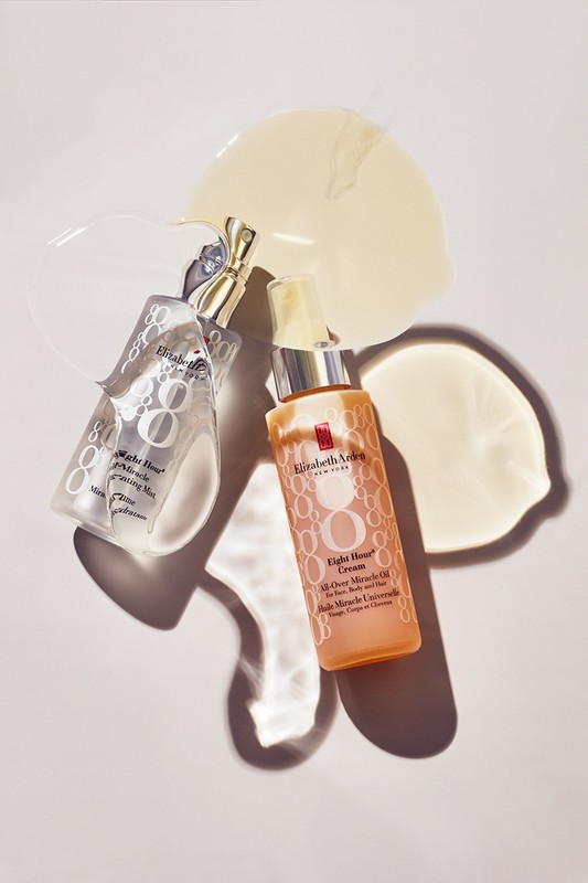 Two Elizabeth Arden skincare bottles sit on a white surface with smears of moisturizer, showcasing beauty product photography with soft lighting.