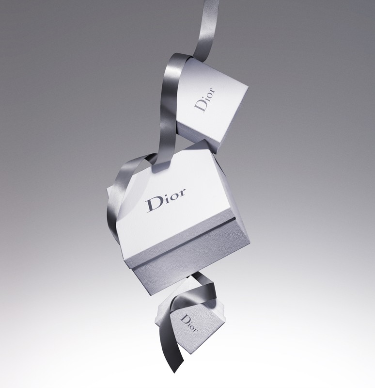 Three Dior branded gift boxes cascade down in a studio shot, connected by a luxurious gray ribbon against a gradient gray background.