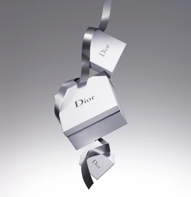 Three Dior branded gift boxes cascade down in a studio shot, connected by a luxurious gray ribbon against a gradient gray background.
