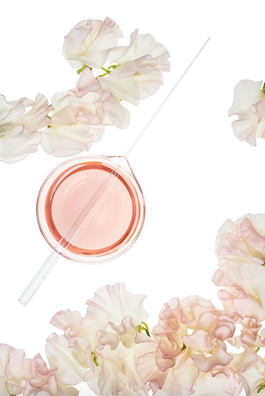 Laboratory beaker filled with pink liquid and a pipette, surrounded by white sweet pea flowers on a white background in a studio shot.