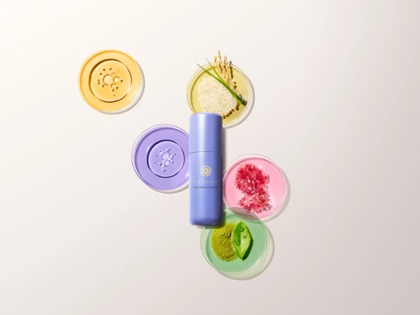 TATCHA The Dewy Serum bottle surrounded by petri dishes with skincare ingredients on a light background. Beauty product shot.
