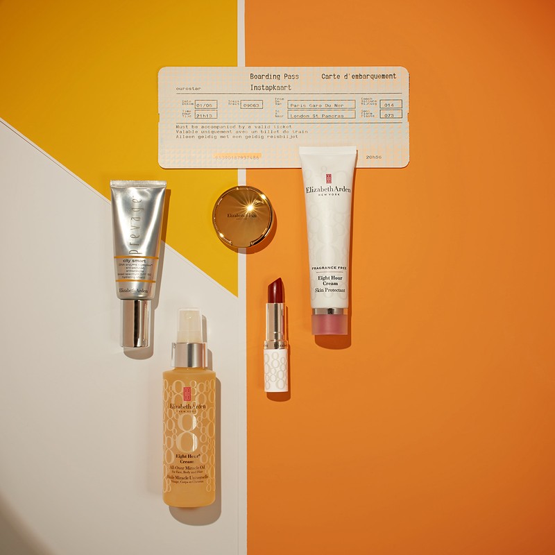 Cosmetic product still life: Elizabeth Arden skincare products, including cream, lipstick, and serum, are arranged on a split orange and white background.
