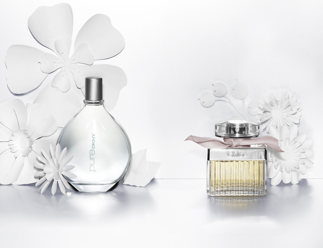Two perfume bottles, one DKNY Pure and one Chloe, are positioned side-by-side against a clean white backdrop decorated with paper flowers.