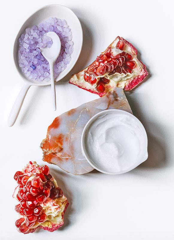 Overhead shot features skincare cream, bath salts, sliced pomegranate, and a decorative stone, creating an organic spa-like product display.